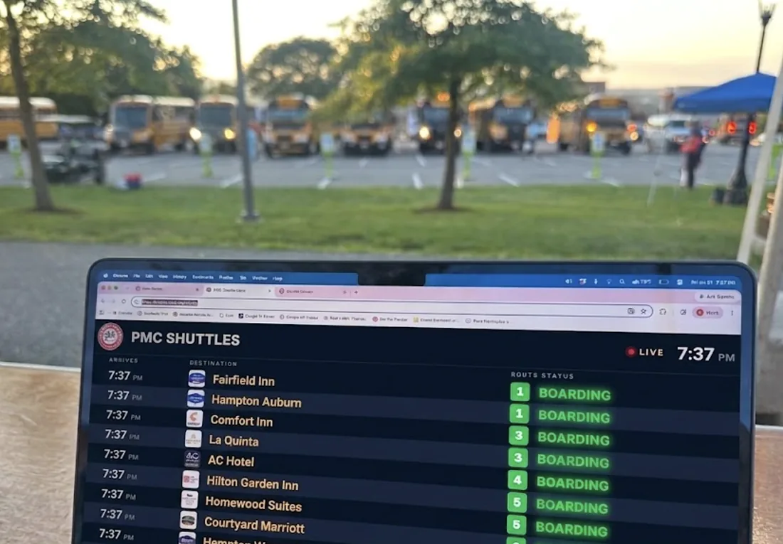 The departures board running on a laptop at the venue at dusk, with the shuttle fleet staged in the lot behind it: Fairfield Inn, Hampton Auburn, Comfort Inn, La Quinta, AC Hotel and more, every one of them boarding at 7:37 p.m.